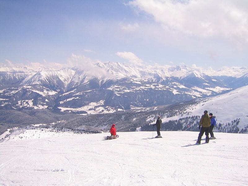 Two skiers on a groomed run with a wide mountain panorama behind