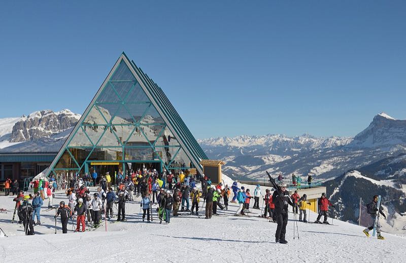 Skiers gathered on the snow outside the glass pyramid of the Boè gondola top station, with Dolomite ridges behind