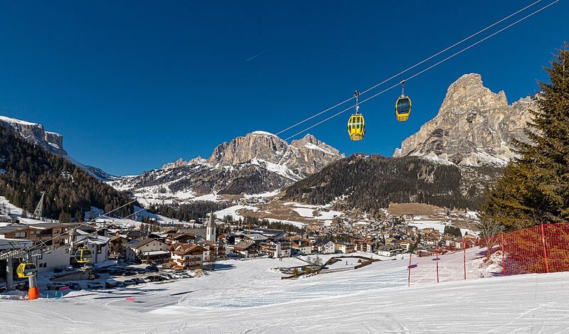 Yellow gondola cabins crossing above a groomed piste, with the village of Corvara and the Sassongher peak under a clear sky