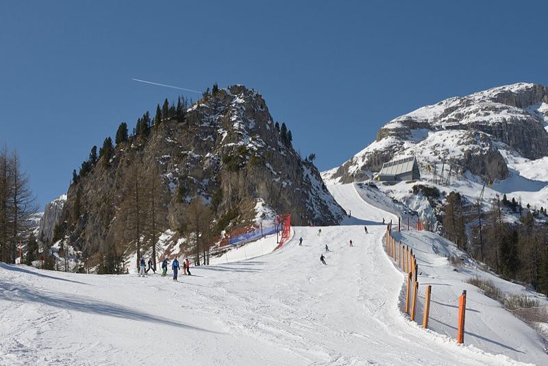 Skiers on a wide groomed piste lined with orange safety posts, below a rocky knoll and a lift station under the Sella cliffs