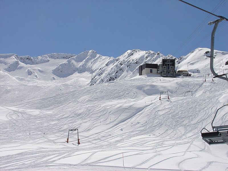 A wide snowfield criss-crossed with ski tracks below a chairlift and a mountain station