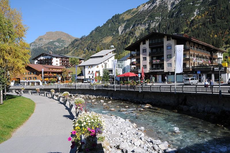 Riverside promenade with flowers in Lech, chalet hotels and a mountain ridge behind under blue sky