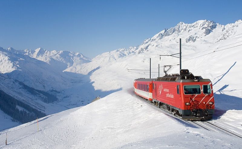 A red mountain train climbing through deep snow above the Urseren valley