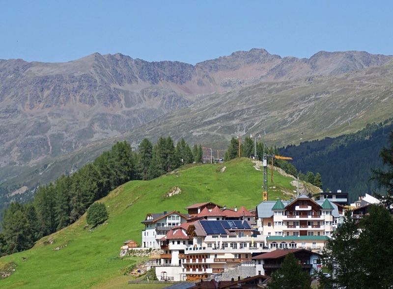 Cluster of Obergurgl chalet hotels on a green alpine hillside below a high mountain ridge under blue sky