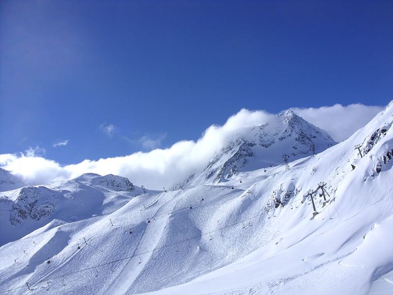 Groomed glacier ski slopes on the Stubai Glacier with the Schaufelspitze wreathed in cloud and lift lines under a deep blue sky