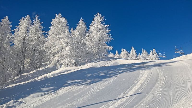 A freshly groomed ski run beside larch trees heavy with frost under a blue sky