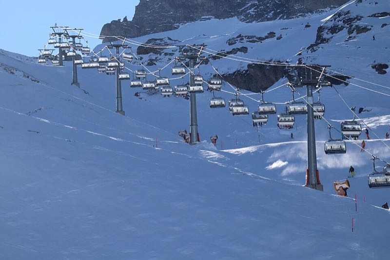 Skiers on a wide piste beneath a chairlift, with snow-covered rock walls behind