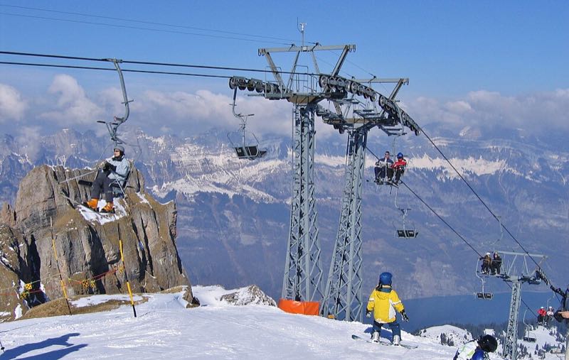 Skiers riding a chairlift past a rock outcrop, with snowy peaks and the lake below