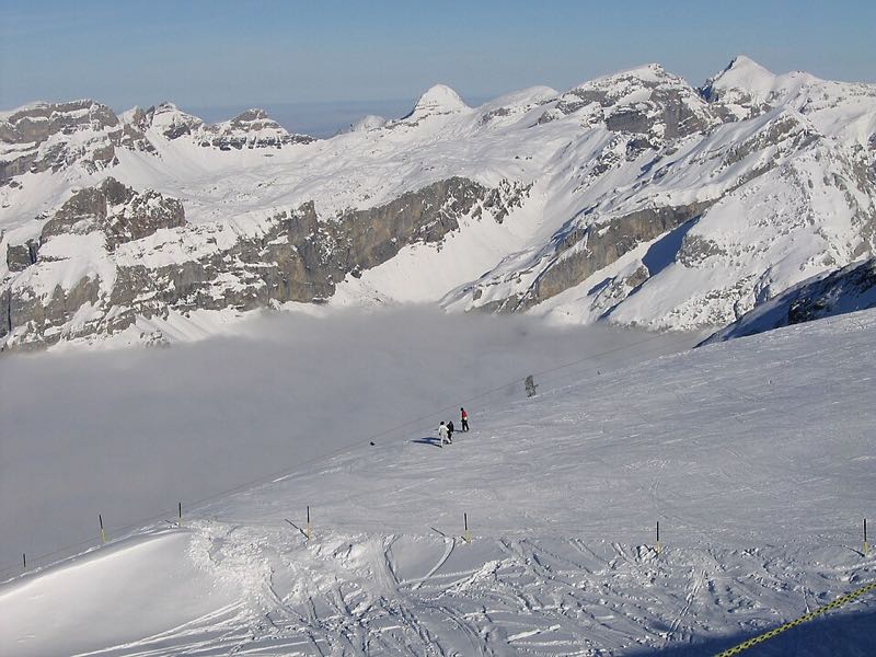 Skiers on a broad glacier piste above a sea of cloud, with limestone peaks across the valley